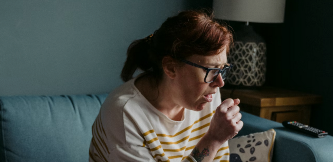 Middle-aged woman coughing in her fist while sitting on her couch at home